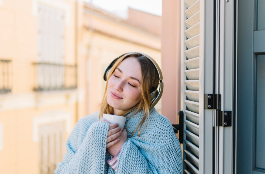 Relaxing everyday sounds shown through a woman listening with headphones by an open window.